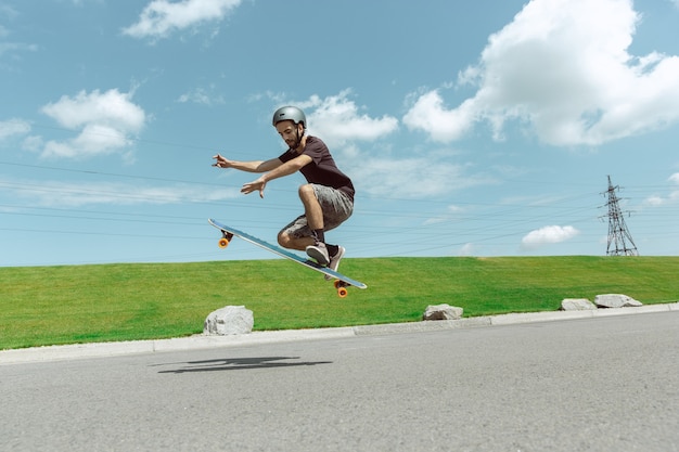 Skater haciendo un truco en la calle de la ciudad en un día soleado. Joven en equipo de equitación y longboard cerca de pradera en acción. Concepto de actividad de ocio, deporte, extremo, afición y movimiento.