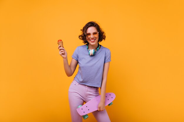 Skater mujer emocionada comiendo helado. Foto interior de niña rizada extasiada con longboard morado.