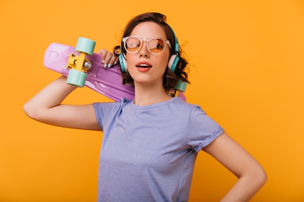 Skater mujer romántica en gafas de moda posando. Foto de chica blanca de moda con longboard morado aislado.