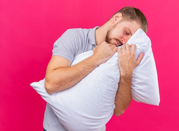 Sleepy joven apuesto enfermo eslavo abrazando la almohada poniendo la cabeza sobre ella con los ojos cerrados aislado sobre fondo carmesí