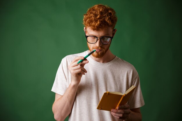 Smart readhead barbudo estudiante en gafas leyendo las notas con bolígrafo en la boca
