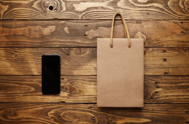 smarthpone negro acostado junto a una bolsa de papel artesanal con asas de cuerda de color marrón claro en una hermosa mesa de madera, tomada desde arriba