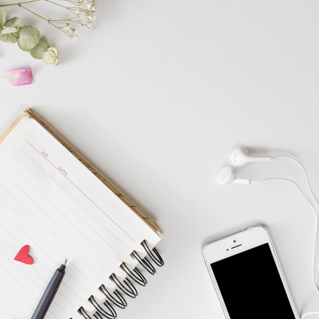 Smartphone con auriculares cerca de cuaderno, pluma, pétalos de rosa fresca y ramitas de plantas