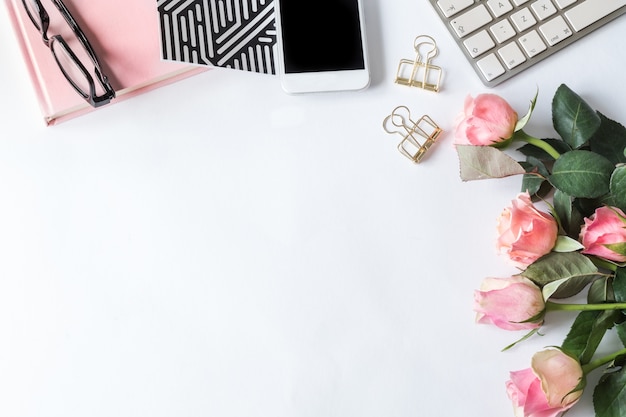 Smartphone, un cuaderno, un teclado, gafas y rosas rosadas sobre una superficie blanca