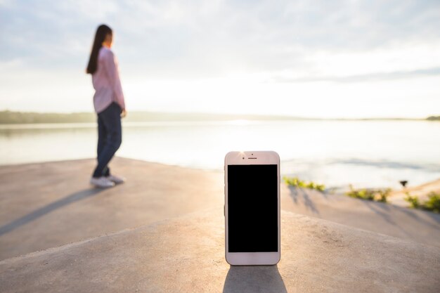 Smartphone frente a mujer de pie cerca del lago idílico