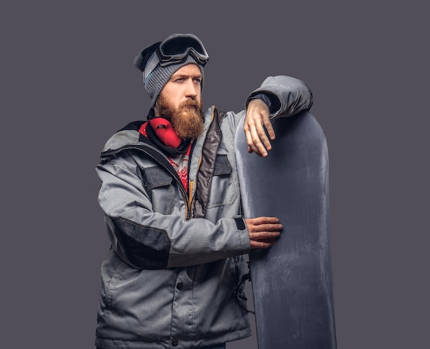 Snowboarder brutal pelirrojo con barba completa en un sombrero de invierno y gafas protectoras vestidas con un abrigo de snowboard posando con snowboard en un estudio, mirando hacia otro lado. Aislado en un fondo gris.