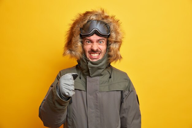 El snowboarder irritado aprieta los dientes por la ira, pasa las vacaciones de invierno en las montañas, aprieta el puño y usa chaqueta y guantes disgustado por el clima frío.