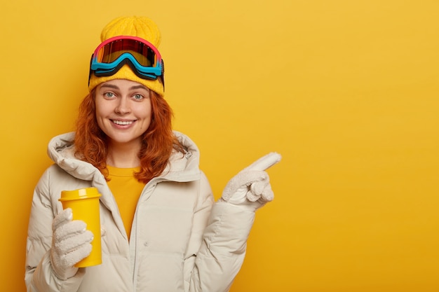 Foto gratuita snowboarder mujer adulta con cabello pelirrojo, disfruta de las bebidas calientes durante el invierno, usa ropa de esquí, señala el espacio libre para su contenido promocional o texto.