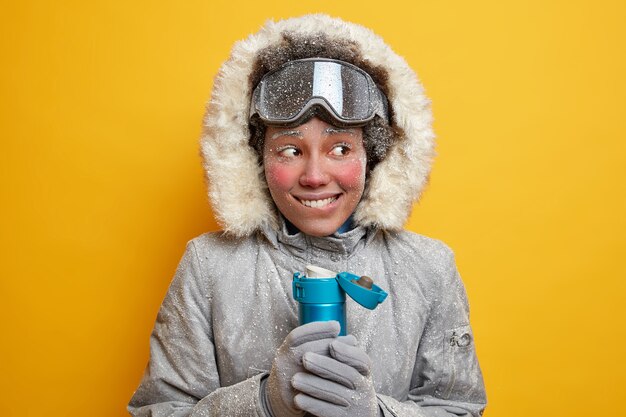 Snowboarder de mujer feliz cubierto con escarcha intenta calentar durante el día helado con bebida caliente vestida con ropa de invierno tiene viaje en el polo norte tiene termo de té. Deporte extremo de temporada