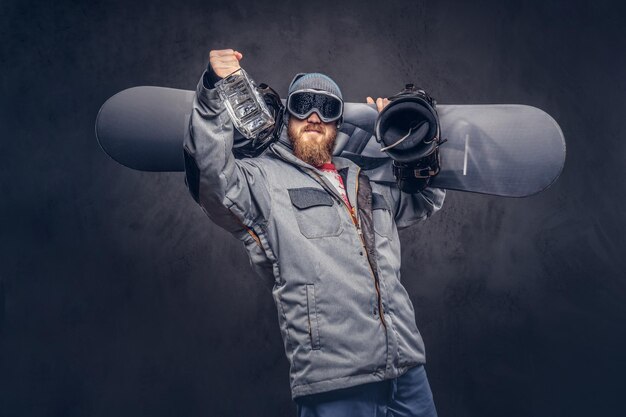 Snowboarder pelirrojo con barba completa en un sombrero de invierno y gafas protectoras vestidas con un abrigo de snowboard sostiene una tabla de snowboard y una botella de alcohol en su hombro en un estudio. Aislado en la espalda gris