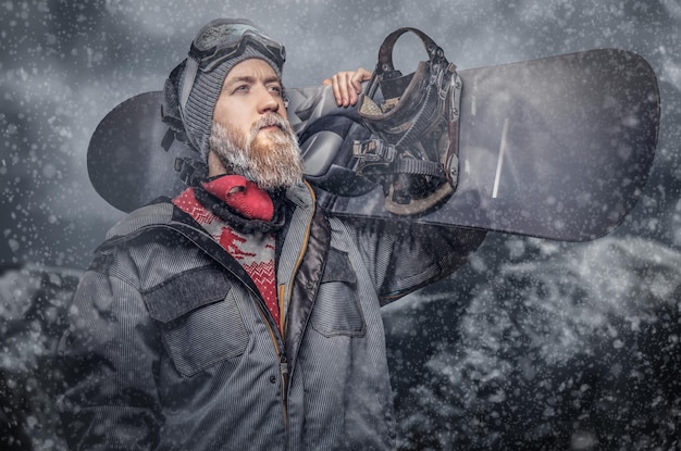 Foto gratuita snowboarder pelirrojo brutal con barba completa en un sombrero de invierno y gafas protectoras vestidas con un abrigo de snowboard posando con snowboard contra el fondo de las montañas.