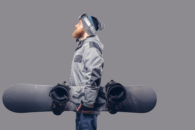 Snowboarder pelirrojo brutal con barba completa en un sombrero de invierno y gafas protectoras vestidas con un abrigo de snowboard posando con snowboard en un estudio, mirando hacia otro lado. Aislado en un fondo gris.