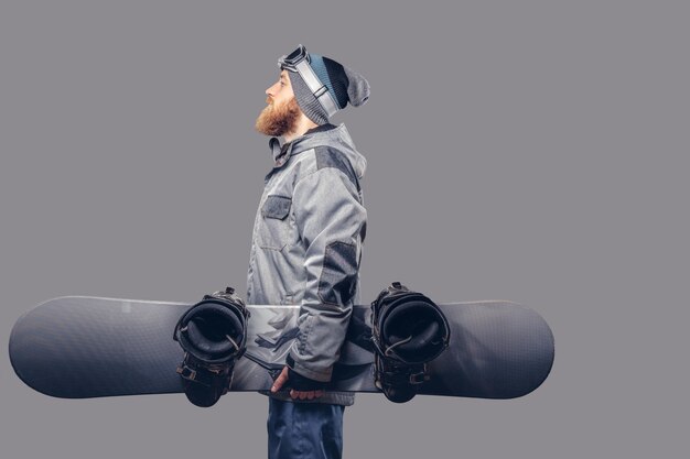 Snowboarder pelirrojo brutal con barba completa en un sombrero de invierno y gafas protectoras vestidas con un abrigo de snowboard posando con snowboard en un estudio, mirando hacia otro lado. Aislado en un fondo gris.