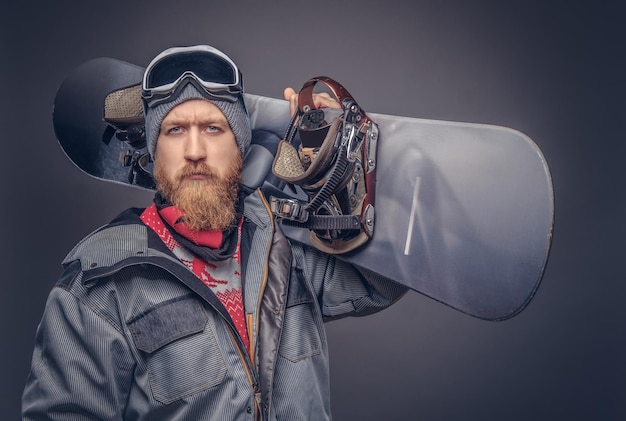 Snowboarder pelirrojo brutal con barba completa en un sombrero de invierno y gafas protectoras vestidas con un abrigo de snowboard posando con una tabla de snowboard en un estudio, mirando una cámara. Aislado en un backgrou gris