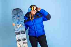 Foto gratuita snowboarding. retrato de mujer caucásica sobre fondo azul de estudio. modelo de mujer hermosa en ropa de abrigo. concepto de emociones, expresión facial, ventas, publicidad. estado de ánimo de invierno, navidad, vacaciones.