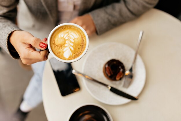 Sobre el bastidor de la mujer joven sostiene una taza de café en el café exterior con postre