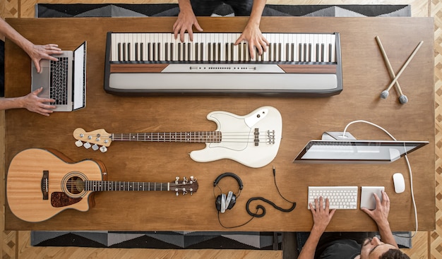 Sobre una mesa de madera hay teclas musicales, guitarra acústica, bajo, mezclador de sonido, auriculares, computadora y baquetas.