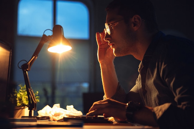 Sobrecargado de trabajo. Hombre trabajando solo en la oficina durante la cuarentena por coronavirus o COVID-19, quedándose hasta tarde en la noche. Joven empresario, gerente haciendo tareas con teléfono inteligente, computadora portátil, tableta en el espacio de trabajo vacío.