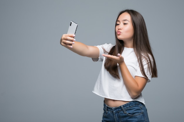 Sociable hermosa chica asiática tomando selfie o hablando en video llamada usando un teléfono celular sobre fondo gris