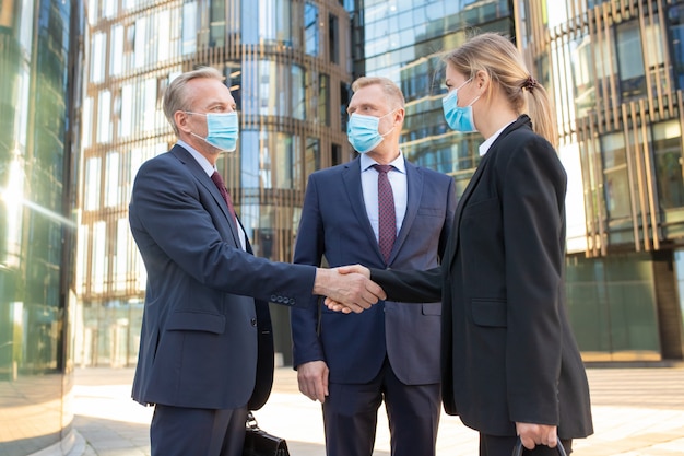 Socios comerciales en máscaras faciales dándose la mano cerca de edificios de oficinas, reuniéndose y hablando en la ciudad. Vista lateral, ángulo bajo. Concepto de comunicación y coronavirus