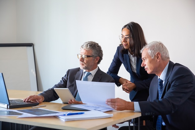 Socios concentrados que usan laptop y trabajan con documentos. Empresarios serios confiados en trajes de oficina discutiendo juntos el proyecto de la empresa. Concepto de gestión, negocios y asociación