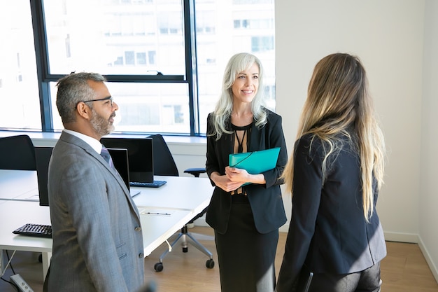 Socios confiados reunidos en la sala de la oficina, hablando y sonriendo. Jefe barbudo en anteojos discutiendo proyecto con hermosas mujeres empresarias. Concepto de negocio, comunicación y alta dirección