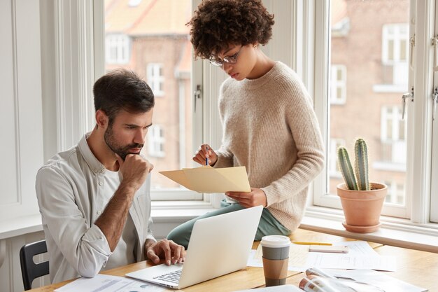 Los socios interraciales trabajan en un proyecto común en el espacio de coworking, discuten ideas, trabajan con papeles, están ocupados, toman café