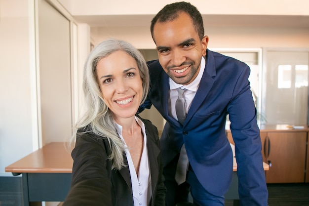 Socios profesionales felices posando para la foto, sonriendo y mirando a la cámara. Empresario de éxito afroamericano y empresaria caucásica tomando selfie. Trabajo en equipo y concepto empresarial