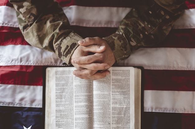 Soldado americano llorando y rezando con la bandera americana y la Biblia delante de él.