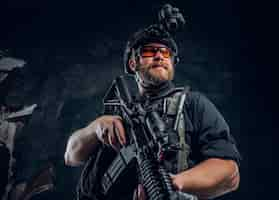 Foto gratuita soldado de las fuerzas especiales barbudo con chaleco antibalas y casco con visión nocturna sosteniendo un rifle de asalto. foto de estudio contra una pared de textura oscura