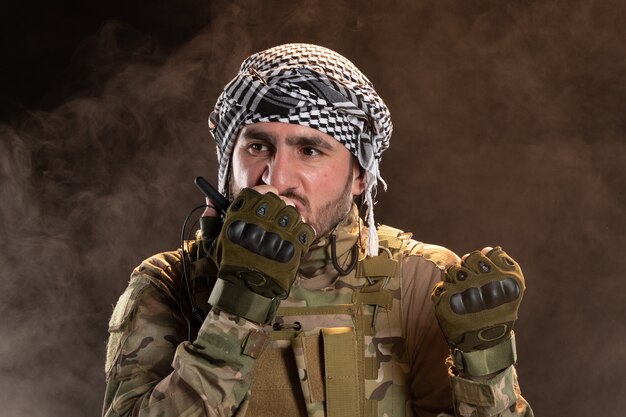 Soldado masculino en camuflaje hablando por radio en pared oscura