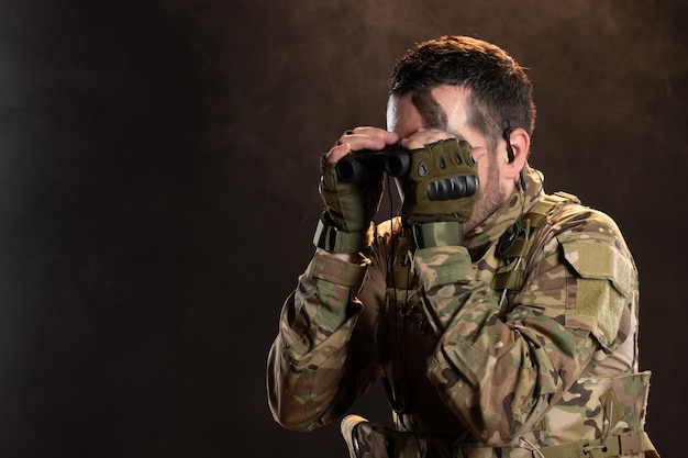 Foto gratuita soldado masculino en uniforme militar mirando a través de binoculares en la pared oscura