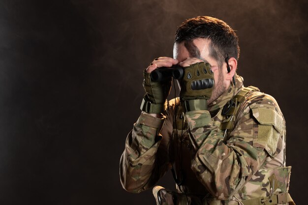 Soldado masculino en uniforme militar mirando a través de binoculares en la pared oscura