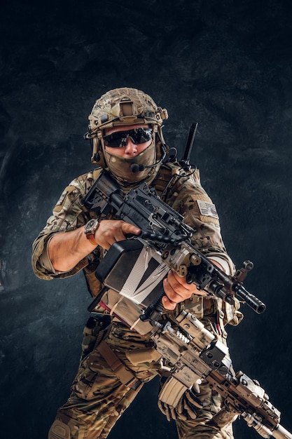 Un soldado serio con equipo completo y uniforme militar está de pie con una ametralladora sobre un fondo oscuro.