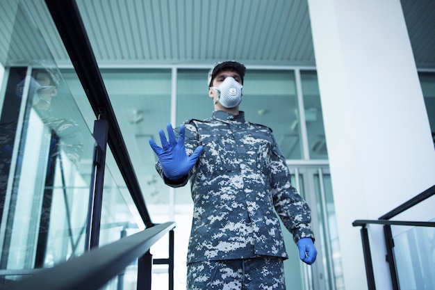 Soldado en uniforme militar con guantes de goma y máscara de protección facial que guarda el hospital y gesticula la señal de stop