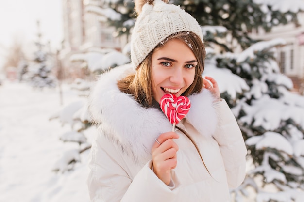 Soleada mañana de invierno en la calle de la encantadora joven lamiendo lollypop rosa. Tiempo feliz, emociones positivas de una mujer bonita en ropa blanca cálida, gorro de punto disfrutando del invierno.