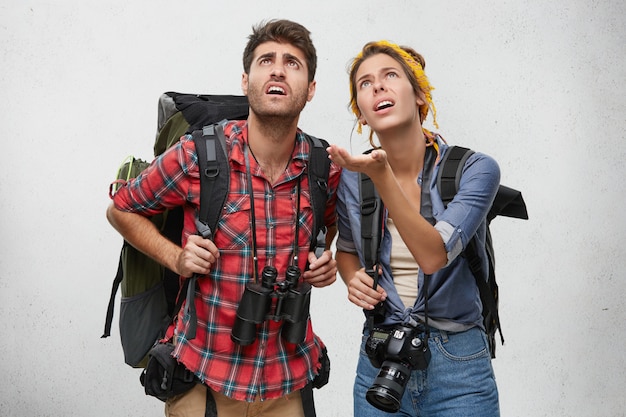 ¡Solo mira esto! Hembra asombrada con la mochila y la cámara que muestran algo con la mano a su marido, siendo sorprendido juntos, aislado. Jóvenes viajeros con mirada sorprendida