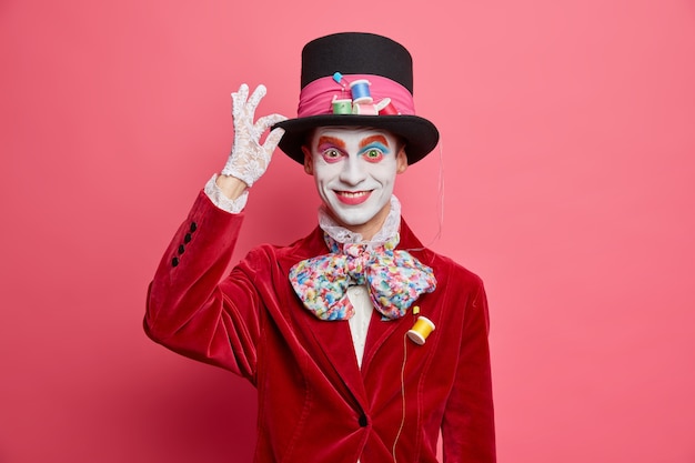 Foto gratuita sombrerero loco positivo mantiene la mano en el sombrero sonríe felizmente participa en vestidos de carnaval para poses de fiesta de halloween contra la pared rosada