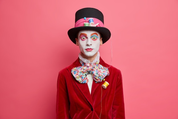 Sombrerero masculino sorprendido usa sombrero pajarita y chaqueta de terciopelo rojo que está presente en el carnaval de halloween usa maquillaje colorido en el interior contra la pared rosada