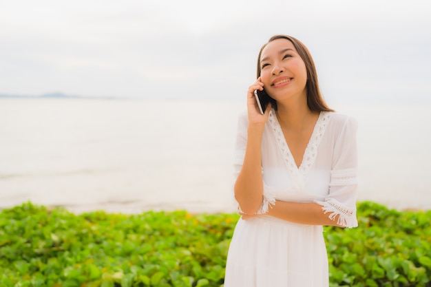 Sombrero asiático hermoso del desgaste de mujer del retrato con la sonrisa feliz para hablar del teléfono móvil en la playa