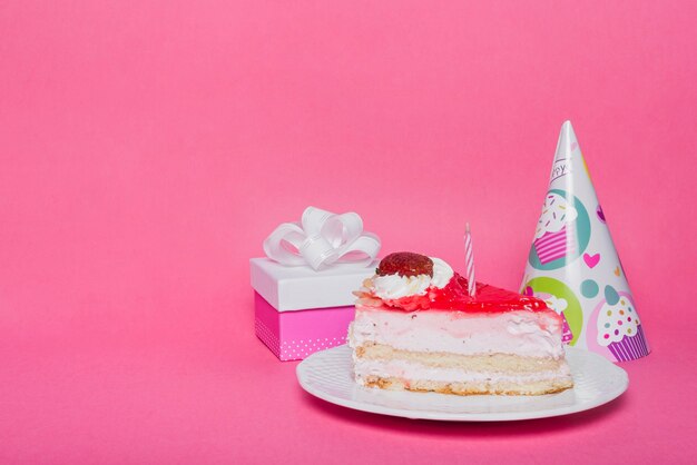 Sombrero de fiesta; caja de regalo y rebanada de pastel con vela sobre fondo rosa