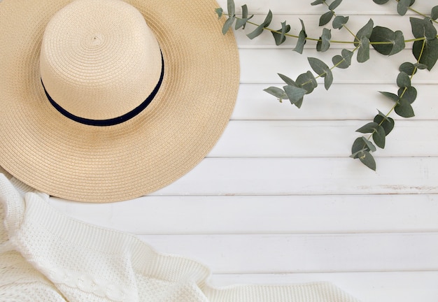 Sombrero de verano y hojas de eucalipto sobre mesa de madera blanca.