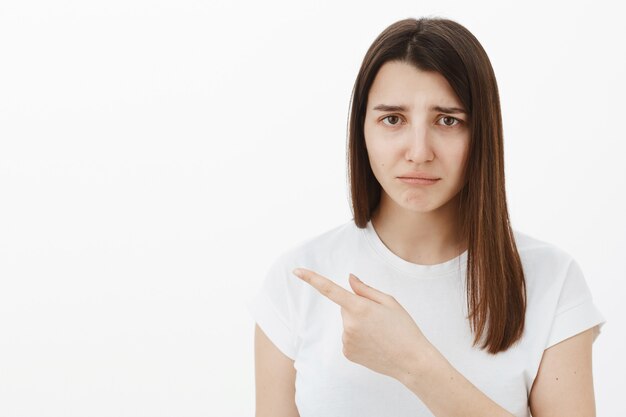 Sombría triste y angustiada linda chica morena con camiseta blanca frunciendo el ceño, frunciendo los labios y apuntando a la izquierda con celos o arrepentimiento, contando lo injusto que sucedió decepcionada y molesta