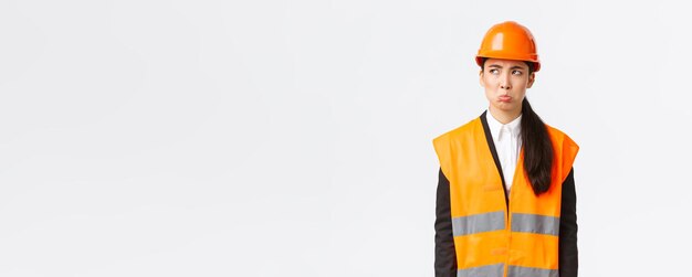 Sombrío y ofendido joven mujer asiática gerente de construcción mujer industrial en casco de seguridad quejándose o lloriqueando haciendo pucheros triste y mirando la esquina superior izquierda decepcionada