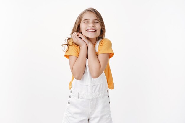 Soñadora alegre niña feliz con cabello rubio, júntese las manos con alegría, no puede esperar un evento excepcional, cierre los ojos sonriendo soñadora, sintiendo alegría y felicidad, coloque la pared blanca