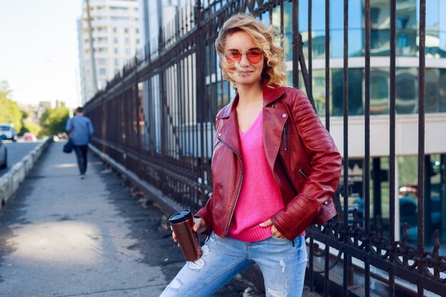 Soñadora chica rubia caminando por las calles, tomando café o capuchino. Elegante traje de otoño, chaqueta de cuero y suéter de punto. Gafas de sol rosas