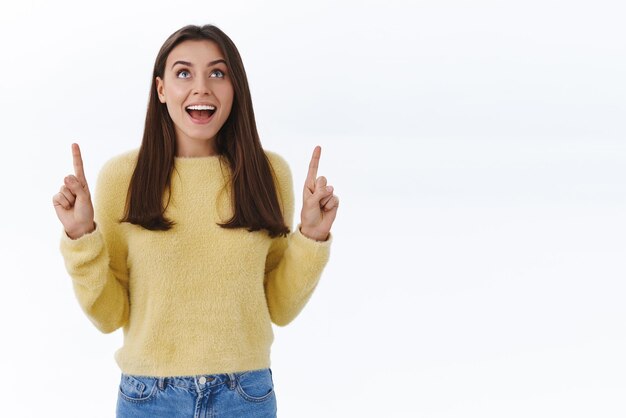 Soñadora y emocionada, la joven morena maravillada se ve fascinada y divertida hacia arriba señalando con el dedo hacia arriba en algo genial e impresionante, disfruta de un hipnótico rendimiento de fuegos artificiales de fondo blanco.