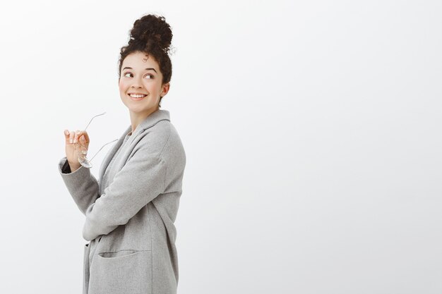 Soñadora empresaria europea urbana de buen aspecto con un elegante abrigo gris, de pie de perfil y mirando a la derecha con una amplia sonrisa segura de sí misma