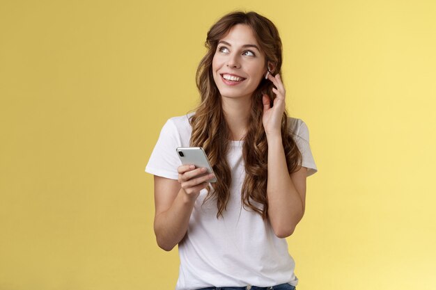 Soñadora feliz alegre niña de pelo rizado mirar a su alrededor contemplar el hermoso clima de verano escuchando música toque auricular inalámbrico llamando amigo hablando a través de auriculares mantenga teléfono inteligente fondo amarillo