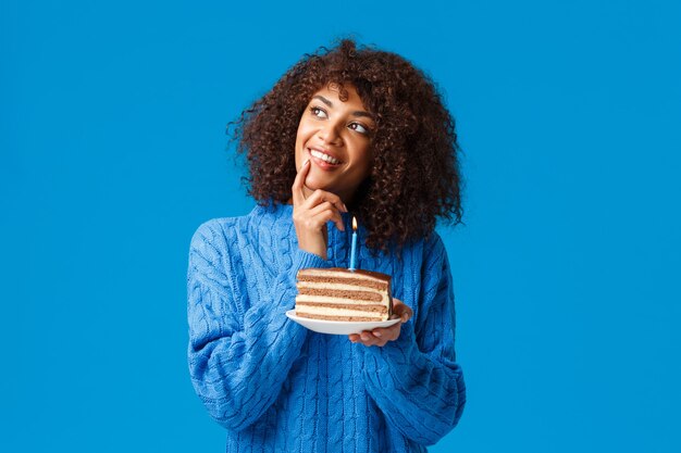 Soñadora y feliz, hermosa mujer afroamericana con corte de pelo afro, pensativa mirando hacia arriba, sonriendo y tocándose el labio como si pensara qué deseo antes de soplar la vela en el pastel de cumpleaños.